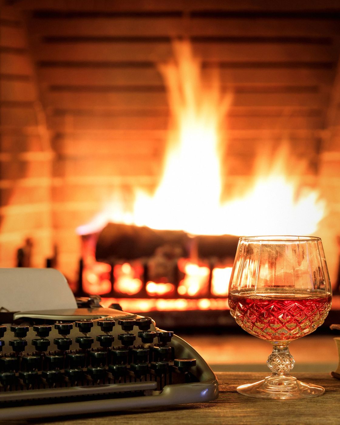 A vintage typewriter, wine glass and book beside a glowing fireplace