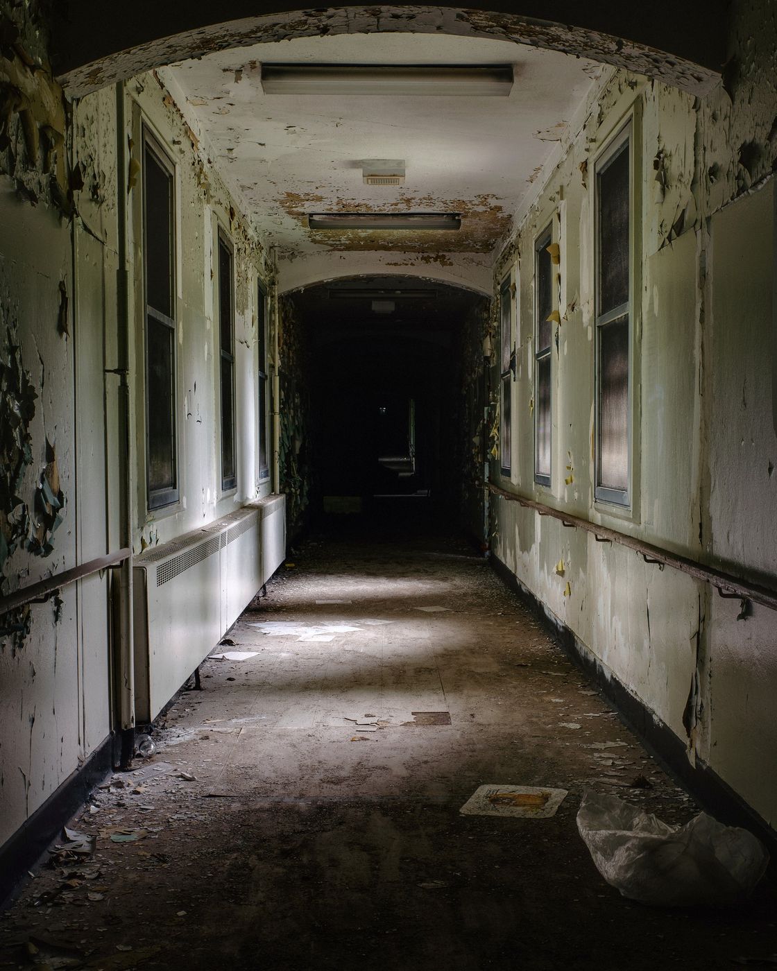 A dark, decaying hallway with peeling walls and a shaft of light at the far end
